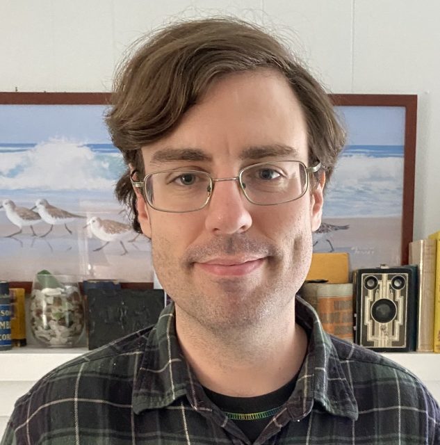 A white, male with brown hair smiling at the camera. Behind him are antique media equipment and a painting of a beach.